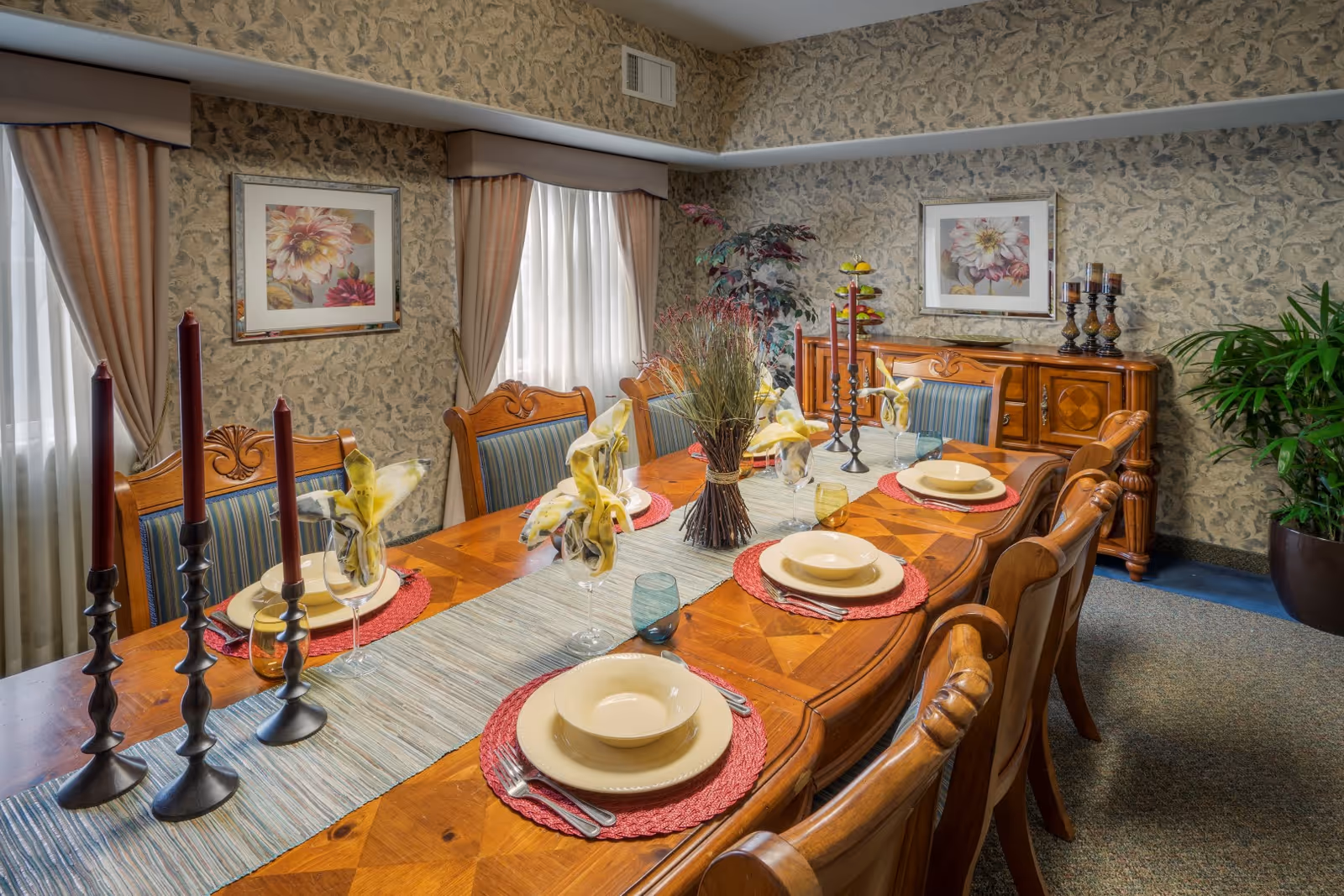 A formal dining room with a long wooden table set for six people. The table has a light blue runner, red placemats, cream-colored plates and bowls, folded yellow napkins in wine glasses, and decorative candlesticks with tall red candles. The room features floral wallpaper, two large windows with beige curtains, framed floral artwork on the walls, a wooden sideboard with candles and a fruit stand, and potted plants in the corners.