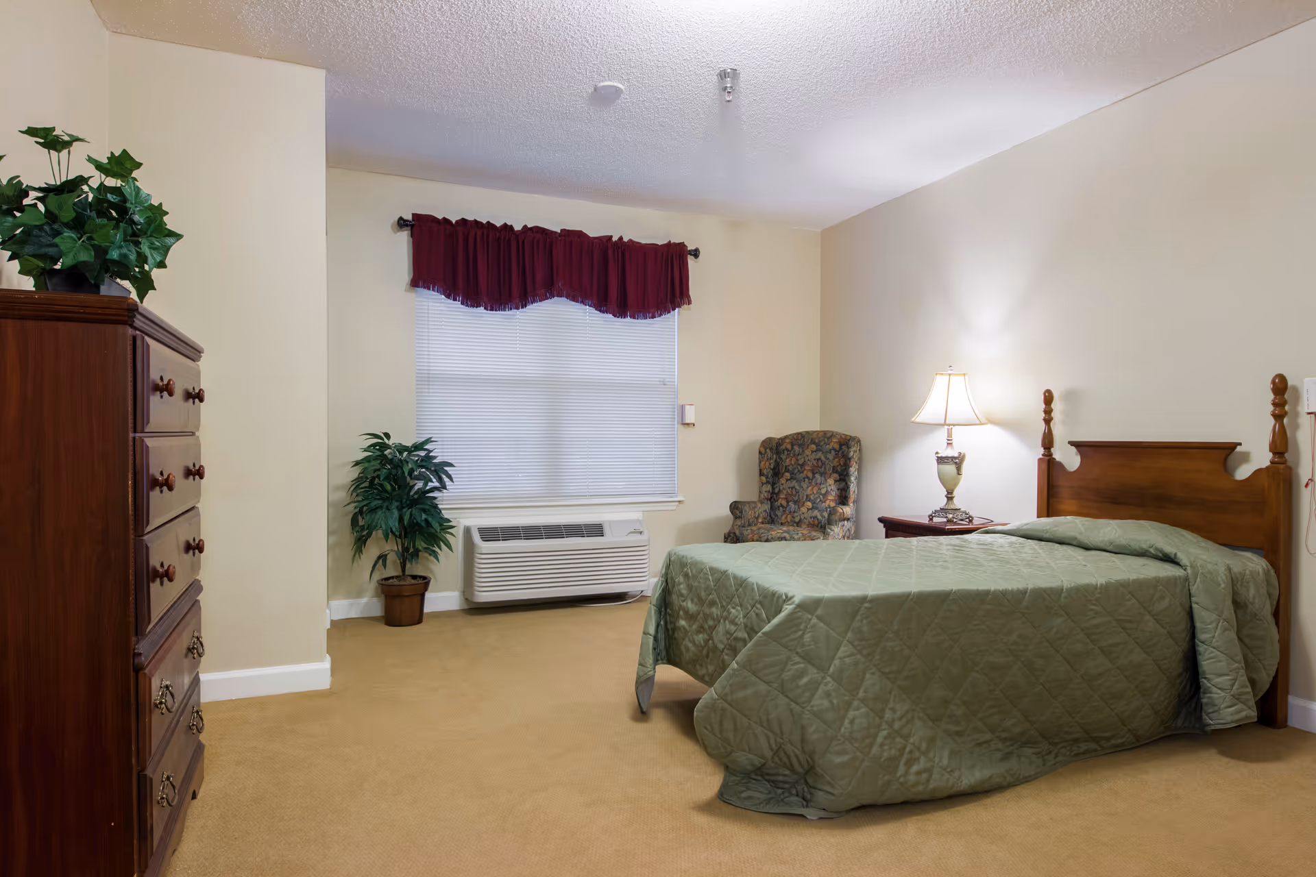 A bedroom with a wooden bed frame and a green quilted bedspread. There is a wooden nightstand with a lamp on it next to the bed. A floral upholstered armchair is placed in the corner near a window with closed blinds and a burgundy valance. A wooden dresser with a potted plant on top is on the left side of the room. The carpet is beige and the walls are light-colored.