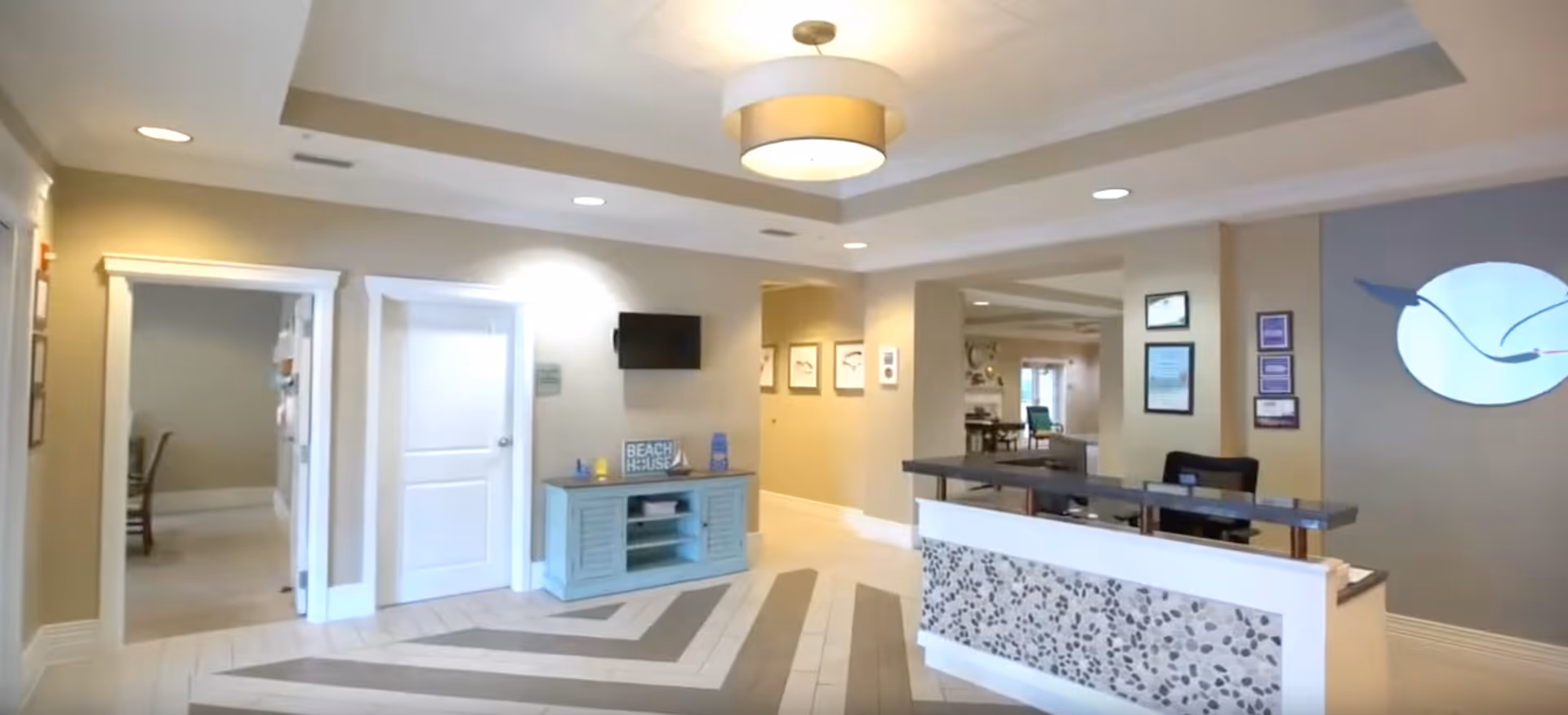 Interior view of a reception area in Beach House Assisted Living & Memory Care featuring a front desk with a decorative stone panel, a light blue cabinet with a small TV mounted above it, and a hallway leading to other rooms. The space is well-lit with recessed lighting and a ceiling light fixture, and the walls are painted in neutral tones.