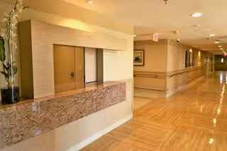 Interior view of a senior living facility hallway with a reception desk on the left side. The hallway has polished wooden floors, beige walls with handrails, ceiling lights, and framed pictures on the walls.