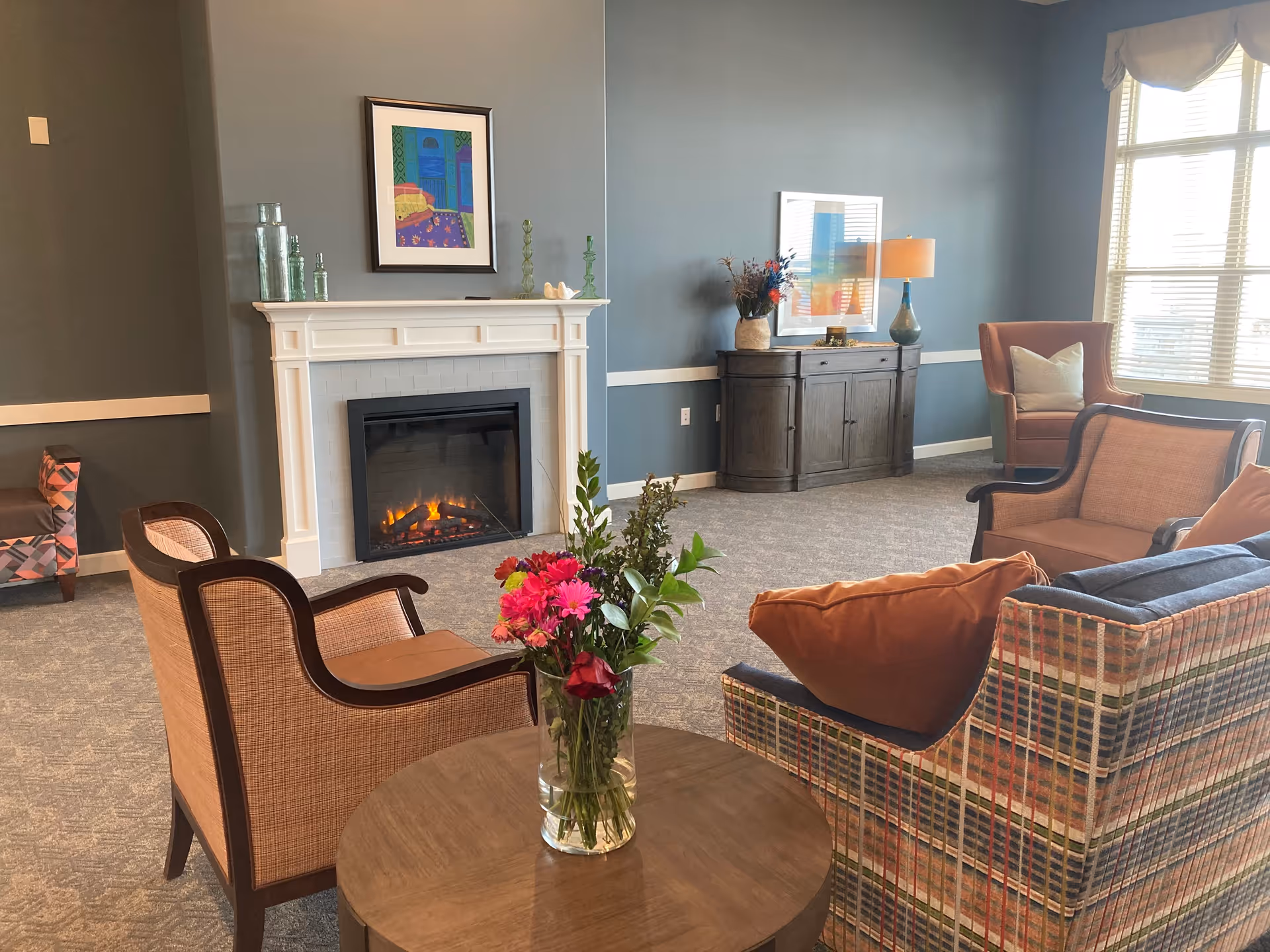 Cozy common room with upholstered chairs around a small table holding a vase of flowers and a lit fireplace against a blue-gray wall.