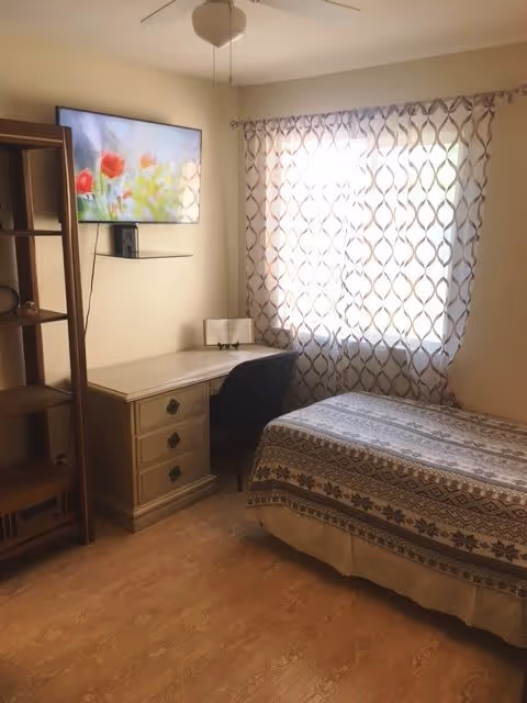 A small bedroom with a single bed covered in a patterned blanket, a wooden desk with drawers, a black chair, a wall-mounted TV displaying a floral image, a wooden shelving unit, and a window with patterned curtains letting in natural light.