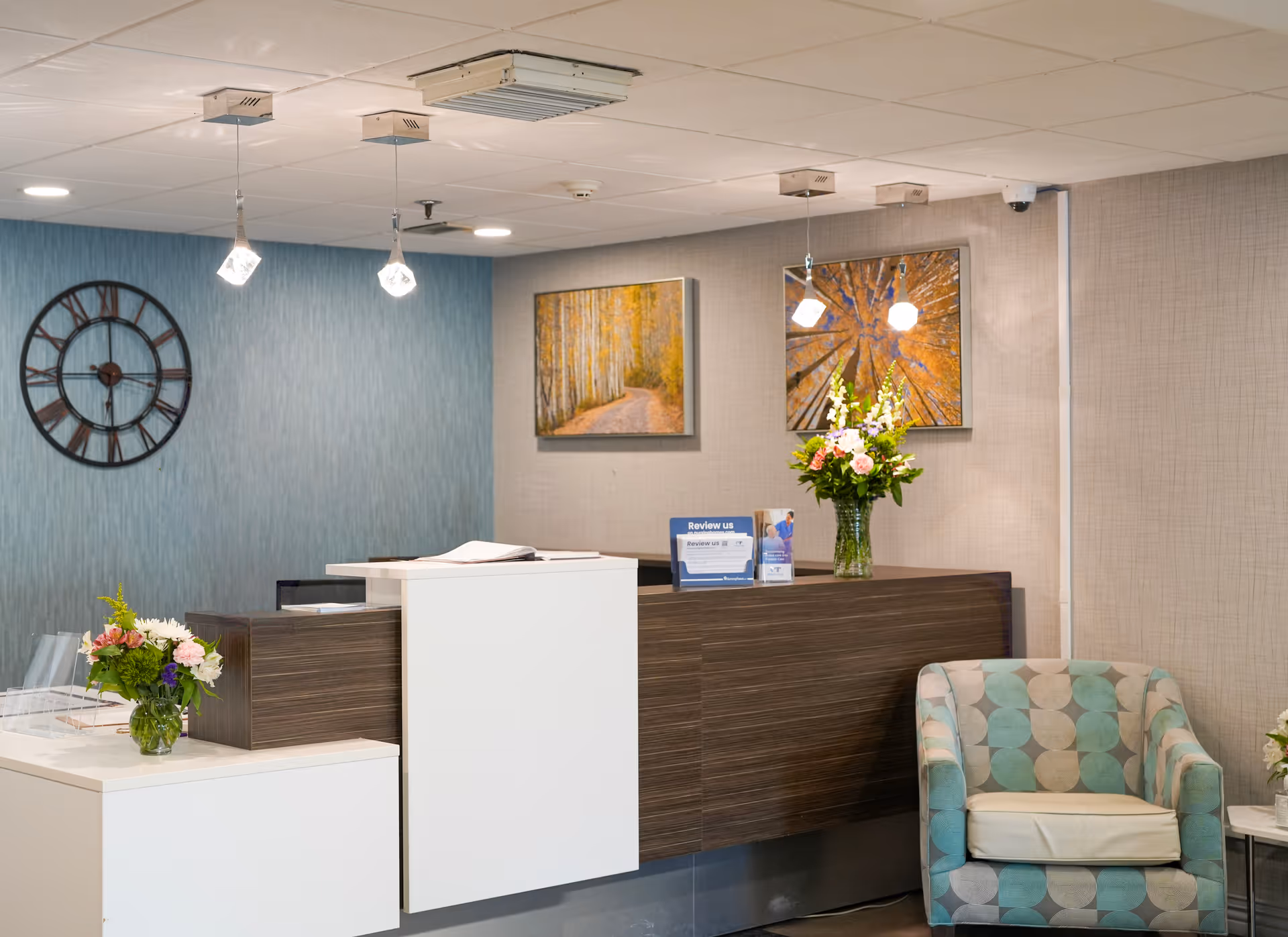 Reception desk in a senior living facility lobby with floral arrangements, wall art, a large clock, and an upholstered chair.