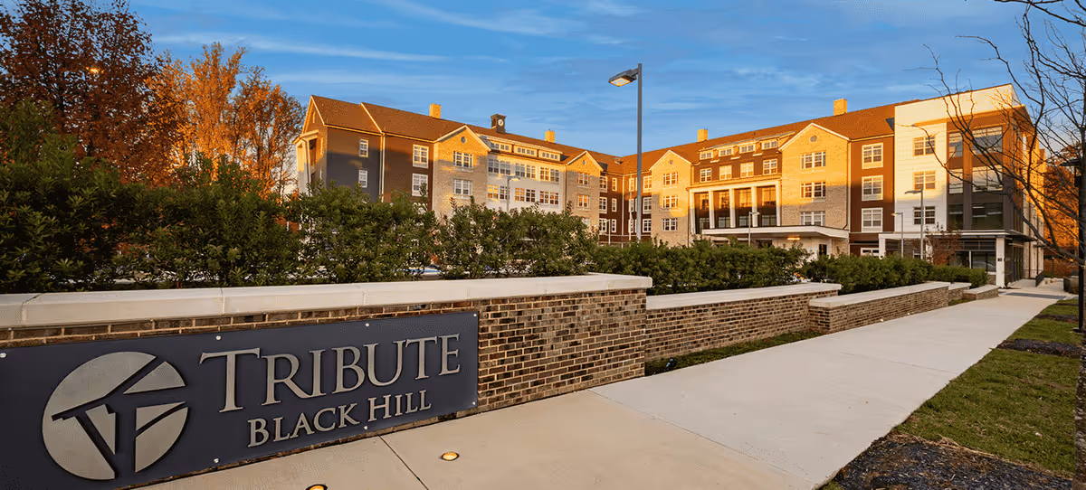 Front exterior of a multi-story senior living building with landscaping and a 'TRIBUTE BLACK HILL' sign in the foreground.