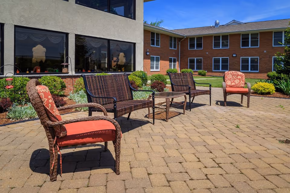 Outdoor patio area at Bentley Assisted Living at Branchville with four wicker chairs and a small round table on a paved surface, surrounded by landscaped bushes and plants, with a two-story brick and stucco building in the background under a clear blue sky.