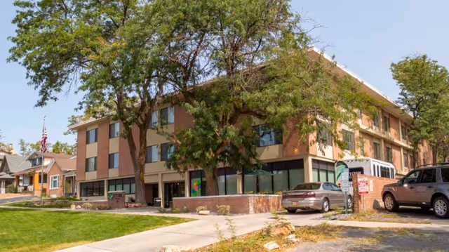 Front exterior of a three-story brick senior living building with trees, parked cars, and a walkway.