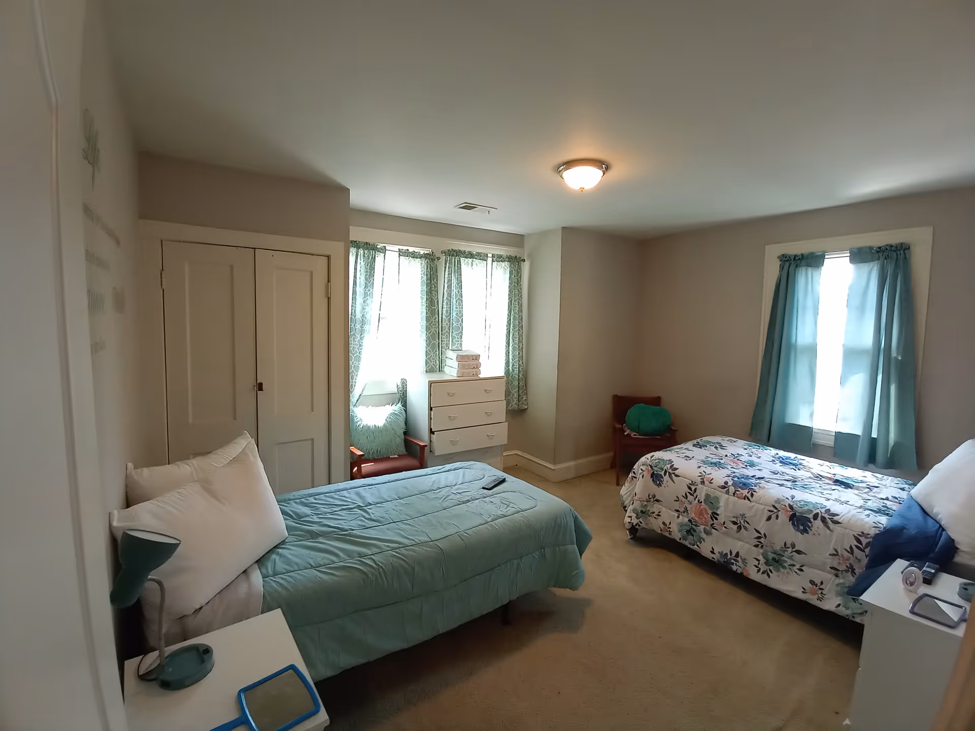Cozy bedroom with two beds, a dresser, chairs, and teal-curtained windows.