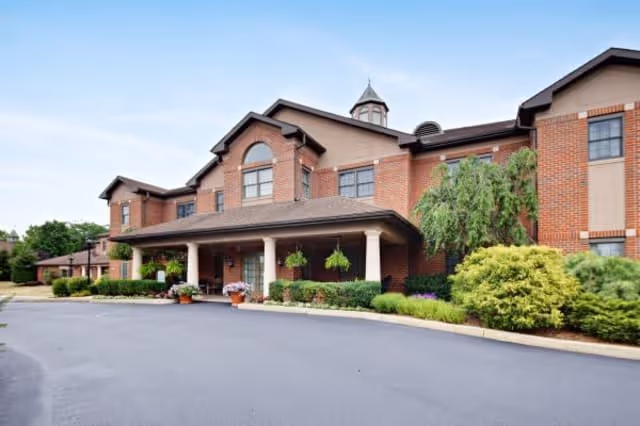 Exterior front view of a two-story senior living facility building with brick and beige siding, a covered entrance supported by columns, hanging plants, and well-maintained landscaping including bushes and trees under a clear blue sky.