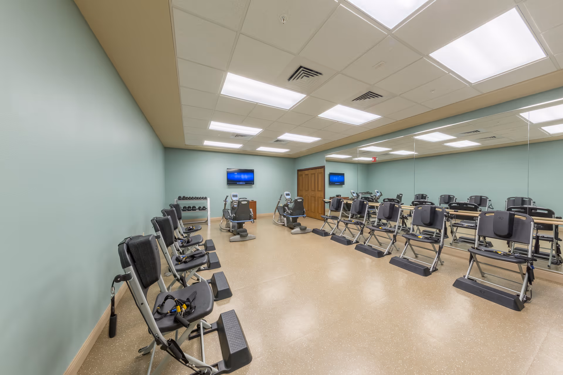 Well-lit exercise room with rows of seated exercise machines, a mirrored wall, dumbbells and a wall-mounted TV.