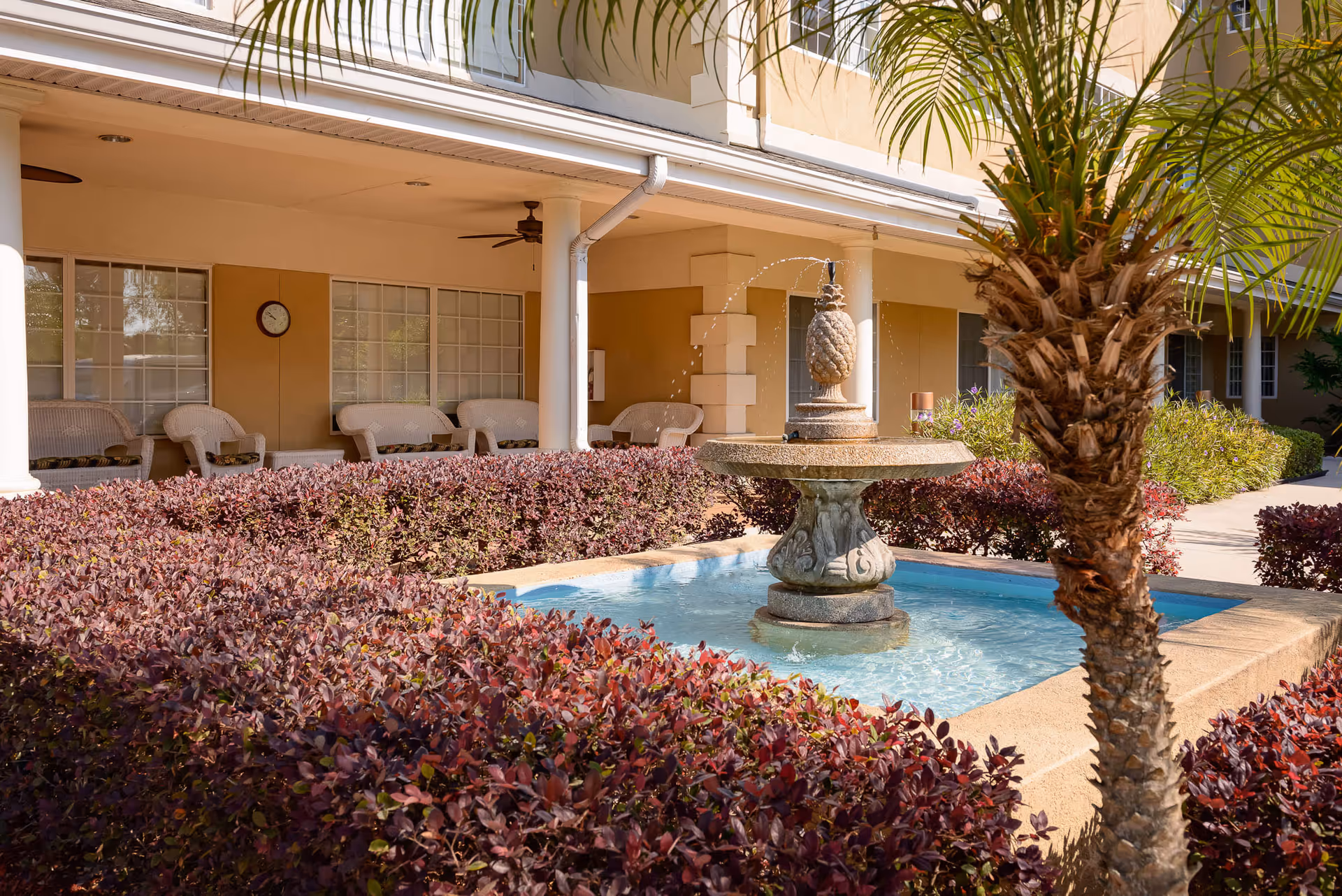Outdoor courtyard area at The Bridge at Ocala featuring a decorative stone fountain with water spouts, surrounded by purple-leaved bushes and a palm tree. In the background, there is a covered patio with white wicker chairs and ceiling fans attached to the building.