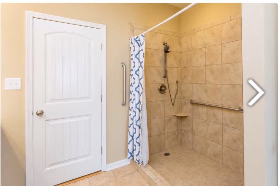 A bathroom shower area with beige tiled walls and floor, a silver showerhead, a grab bar on the wall, and a blue and white patterned shower curtain. There is a white door with a silver knob next to the shower.