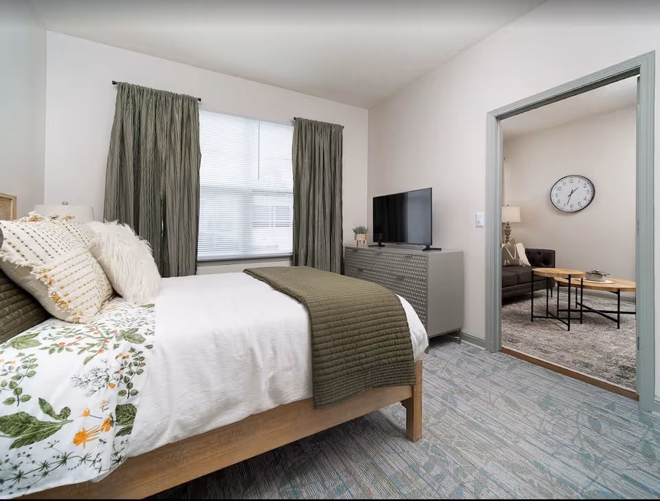 A bedroom with a bed featuring white and floral bedding, green throw blanket, and multiple pillows. There are green curtains covering a window behind the bed. A gray dresser with a flat-screen TV and a small plant is against the wall. Through an open doorway, a living room is visible with a dark sofa, two round wooden coffee tables, a floor lamp, and a wall clock.