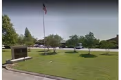 Front lawn and driveway of a senior living facility showing a flagpole with an American flag, a small entrance sign, trees, and parked vehicles.
