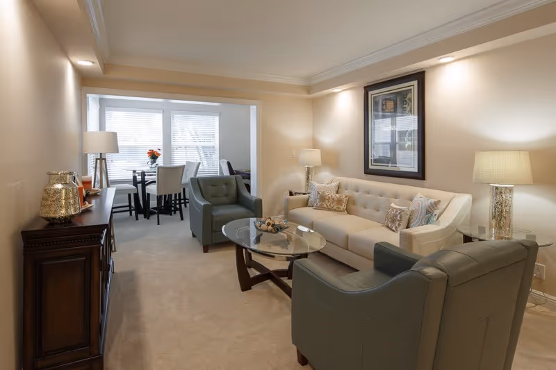 A cozy living room with a beige sofa, two green armchairs, a glass coffee table, and two side tables with lamps. There is a framed artwork on the wall above the sofa. In the background, a dining area with a table and chairs is visible near a window with blinds.