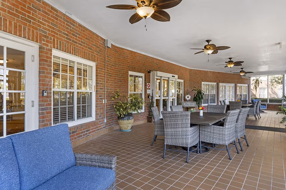Covered outdoor patio area with brick walls, ceiling fans, and multiple seating arrangements including a blue cushioned sofa and gray wicker chairs around tables. Potted plants and large windows are visible along the wall.