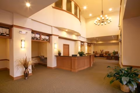 Spacious reception area with a wooden front desk, beige walls, carpeted floor, decorative plants in vases, and a modern chandelier hanging from the ceiling. The area has a high ceiling with a balcony above and seating visible in the background.