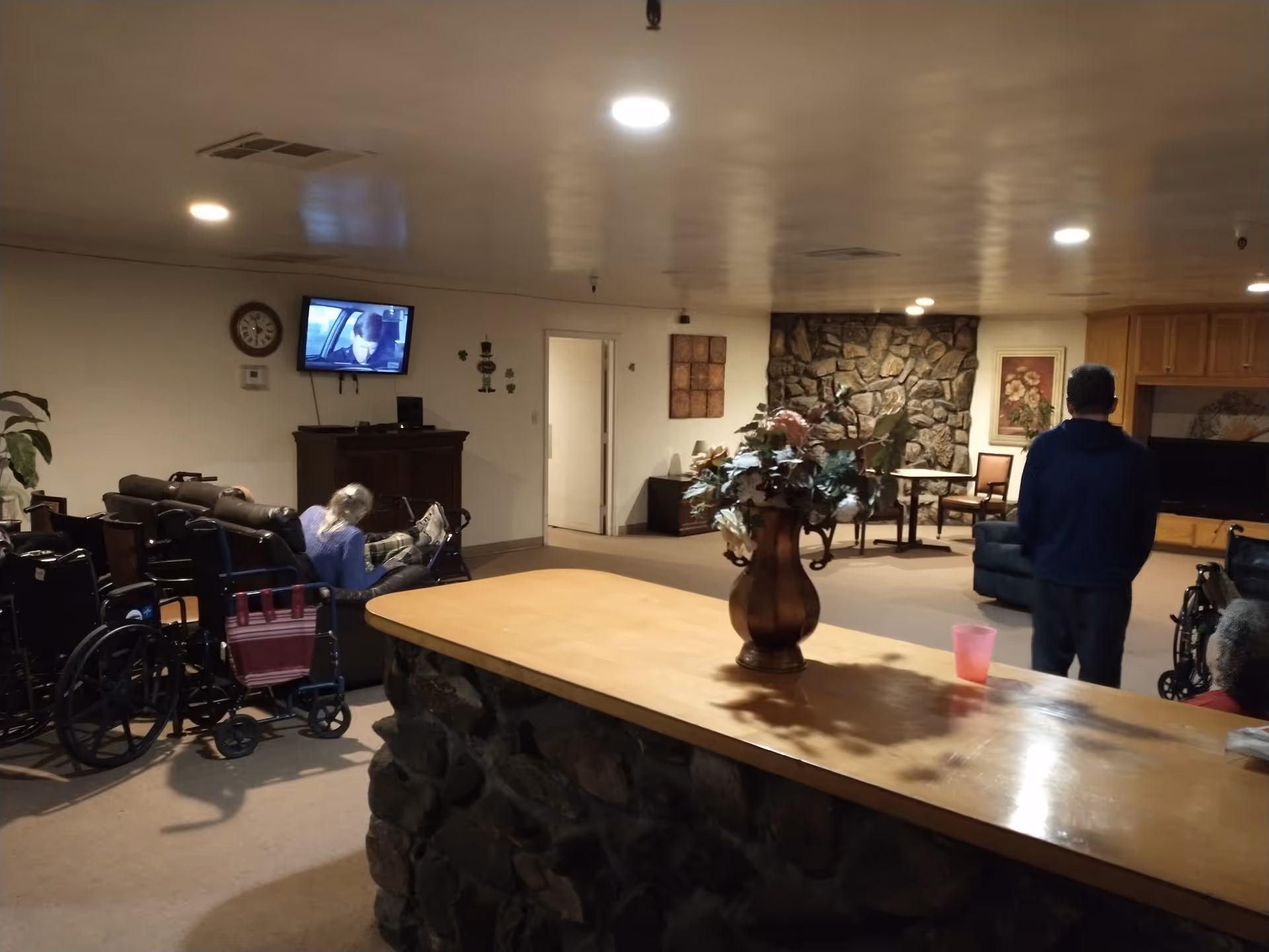 Interior view of a common living area in an assisted living facility with a stone counter in the foreground decorated with a vase of flowers. Several wheelchairs are visible, along with a few elderly residents seated on couches and a man standing. A TV is mounted on the wall showing a person, and the room has warm lighting with stone and wood decor.