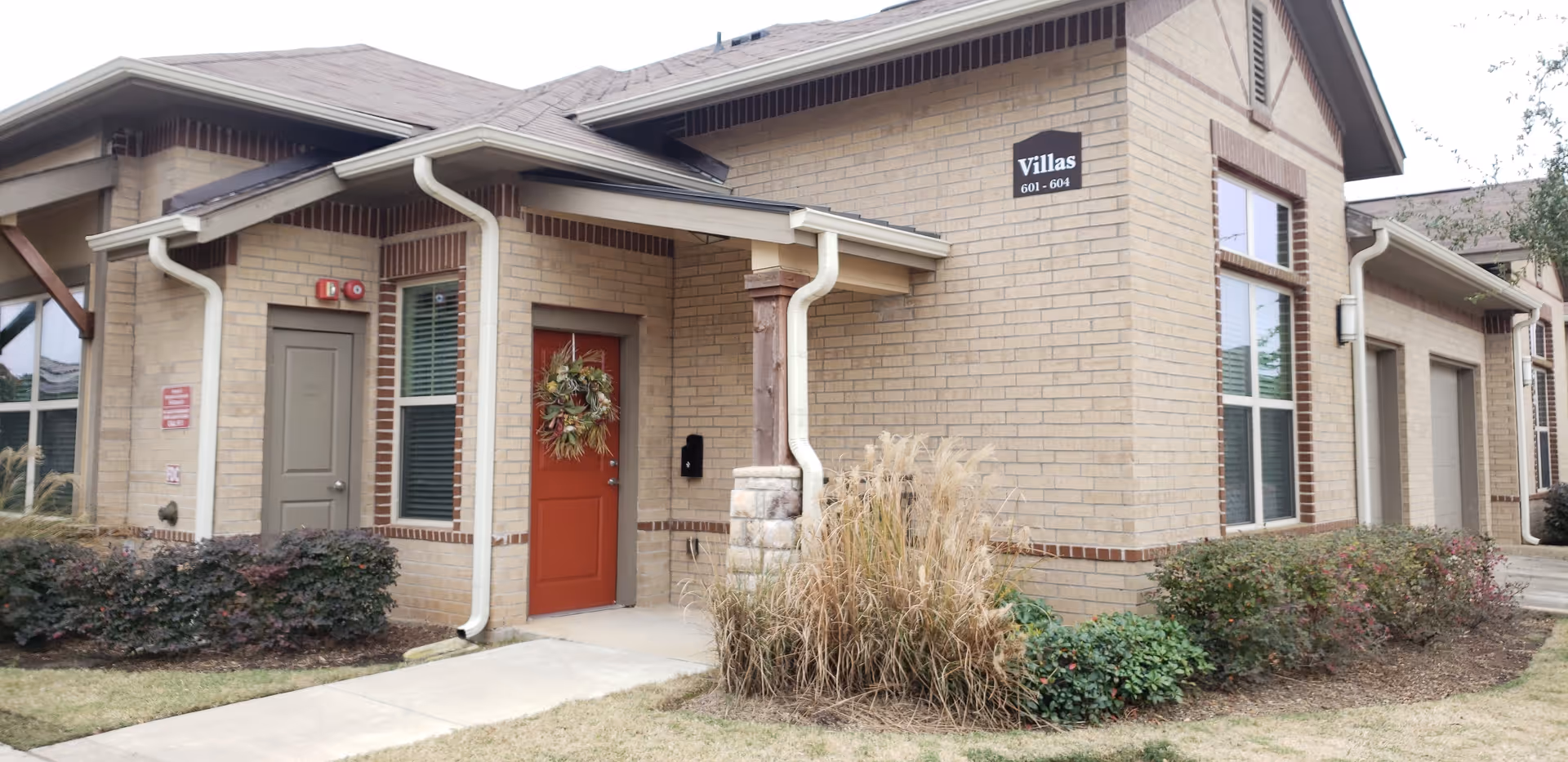 Exterior view of a single-story brick villa unit at Landon Ridge Sugar Land Independent Living with a red front door decorated with a wreath, windows with blinds, and surrounding landscaping including bushes and ornamental grasses.