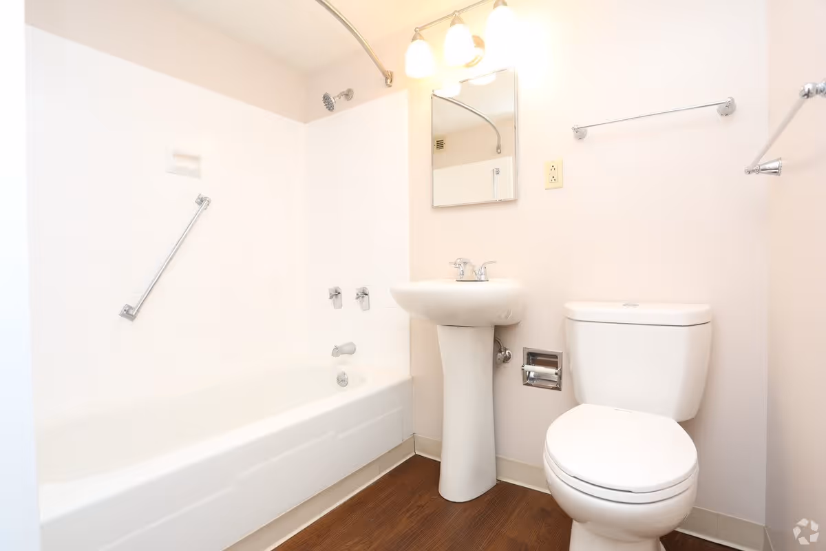 A clean bathroom featuring a white bathtub with a showerhead and grab bar, a white pedestal sink with a mirror above it, and a white toilet. The walls are light-colored and the floor has a wood-like finish.