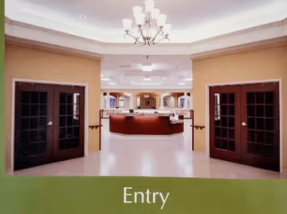 Interior view of an entry area in a facility with double wooden doors on both sides, a chandelier hanging from the ceiling, and a reception desk in the background. The walls are painted beige and the floor is light-colored. The word 'Entry' is displayed at the bottom of the image.