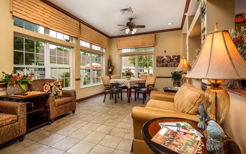 A cozy living room area in a senior living facility with large windows letting in natural light. The room features a tan sofa with a side table holding magazines and a decorative lamp, two patterned armchairs with a small table between them, and a round table with four chairs near the windows. The walls are painted a warm beige, and there are decorative plants and artwork on the walls.