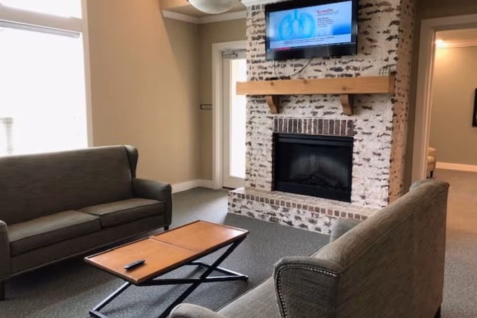 A cozy living room area with two gray upholstered sofas facing each other and a wooden coffee table in between. A brick fireplace with a wooden mantel is centered on the far wall, above which a flat-screen TV is mounted. The room has beige walls, a window letting in natural light, and a door leading outside.