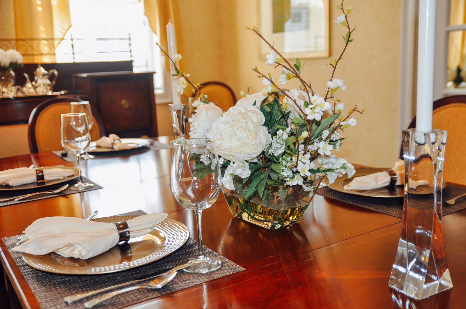 A dining table set for a meal with silver plates, white napkins with napkin rings, wine glasses, silverware, and a floral centerpiece with white flowers and green leaves. The background shows a window with curtains and a wooden sideboard with a silver tea set.