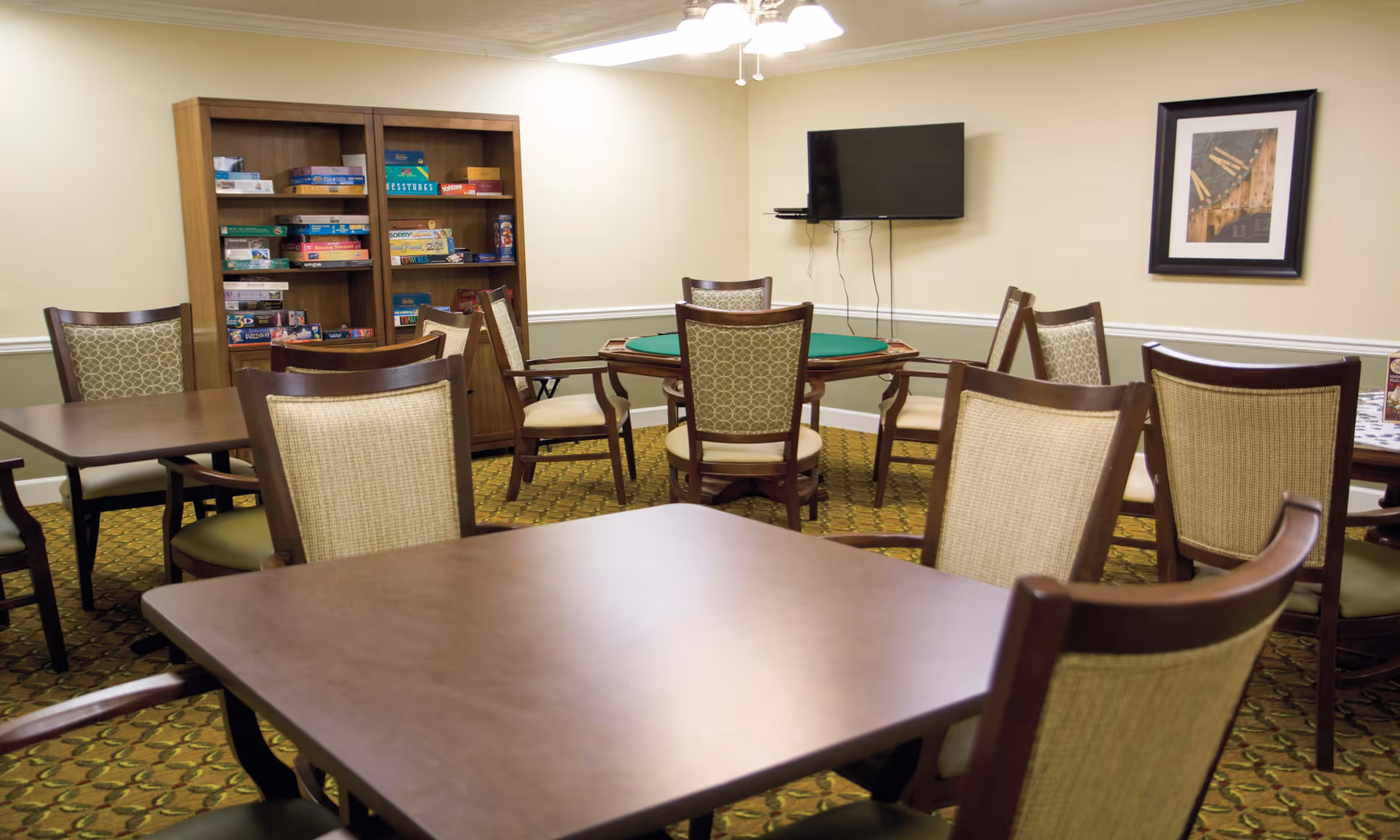A well-lit common room with multiple wooden tables and chairs arranged for group activities. A bookshelf filled with board games is against the back wall. A flat-screen TV is mounted on the wall, and a framed picture hangs nearby. The room has a patterned carpet and light-colored walls with a chair rail.