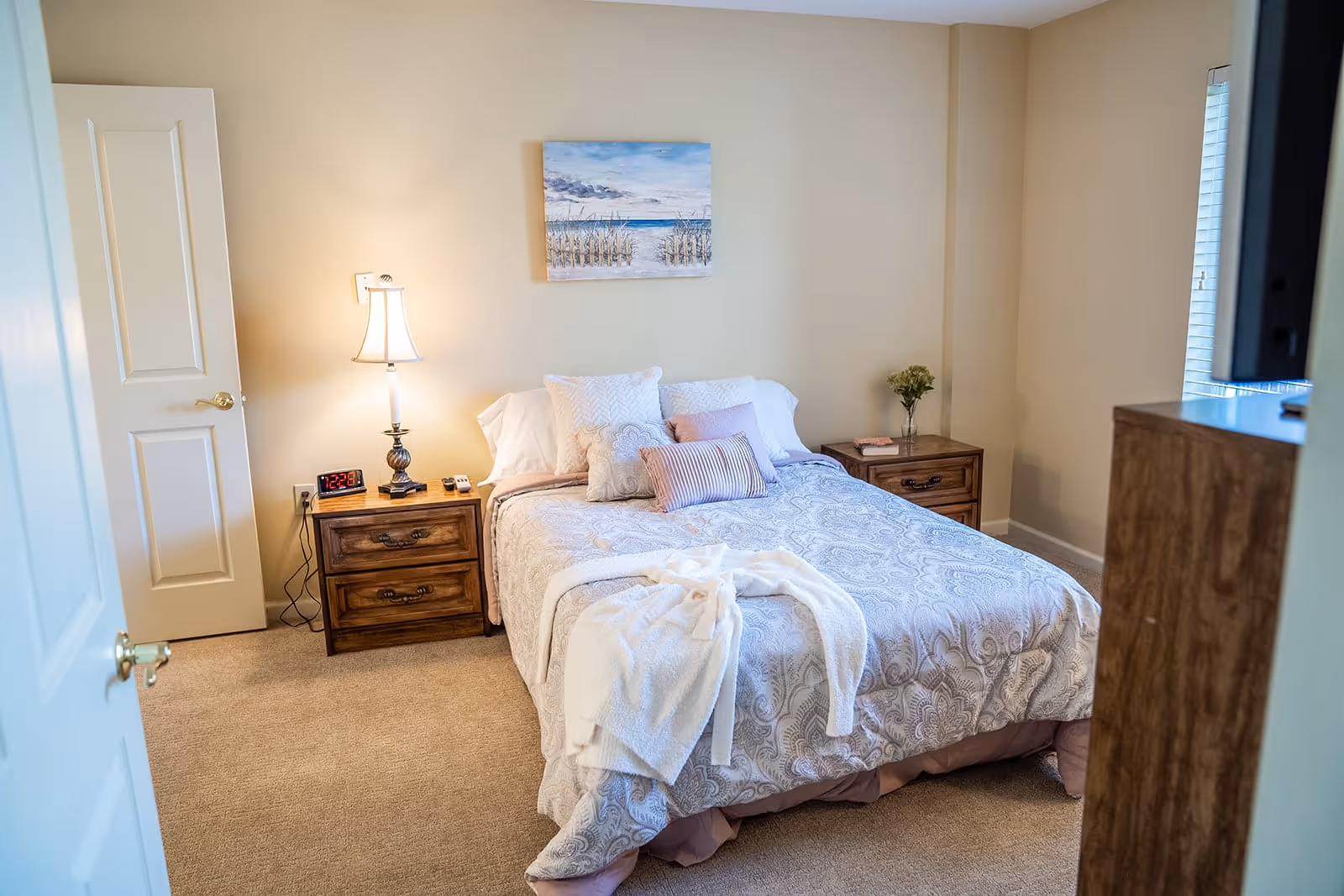 A cozy bedroom with a neatly made bed featuring patterned bedding and several pillows. There are two wooden nightstands on either side of the bed, one with a lamp, alarm clock, and remote controls, and the other with a small vase of flowers and a book. A painting of a beach scene hangs on the wall above the bed. The room has beige walls and carpeted floor, with a partially open door on the left and a window with blinds on the right.