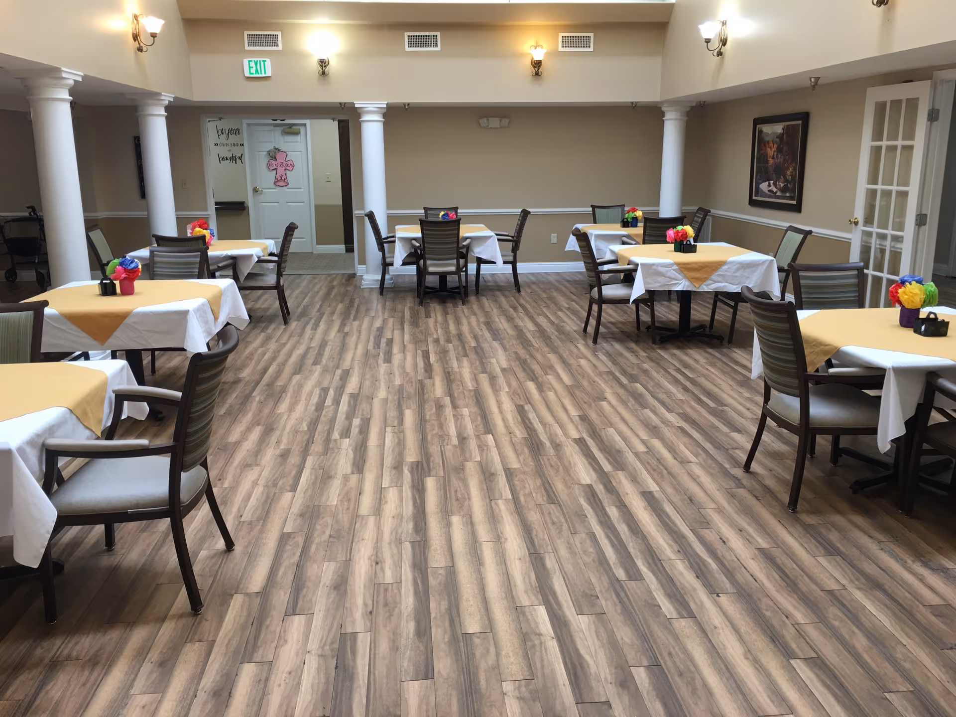 A dining room with several tables covered with white and yellow tablecloths, each decorated with colorful flower centerpieces. The room has wooden flooring, beige walls, white columns, and wall-mounted lights. There is an exit sign above a door at the back of the room.