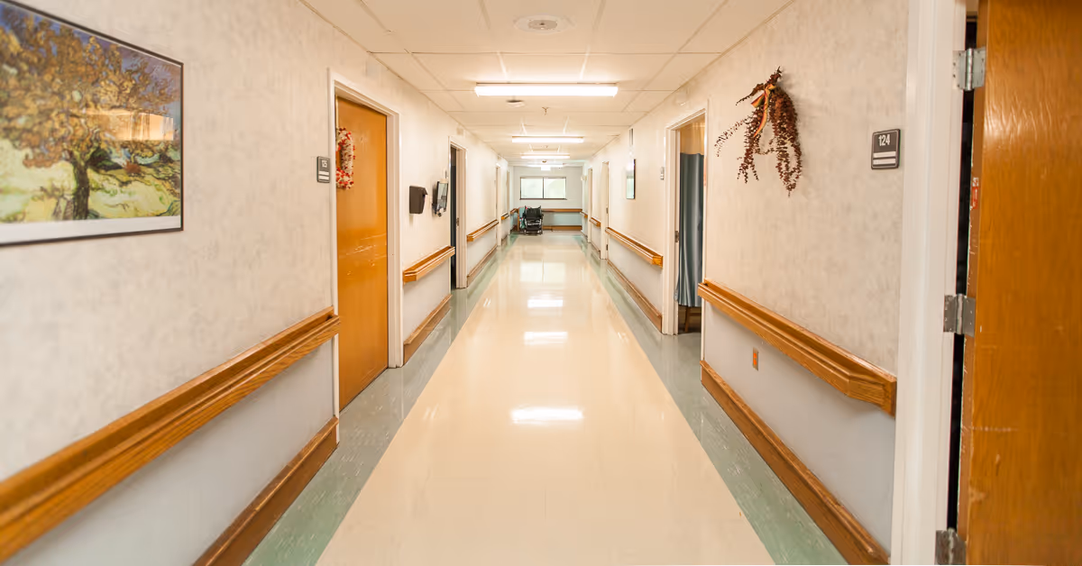 A clean, well-lit hallway in a senior living facility with wooden handrails on both sides, several closed doors, a painting on the left wall, and a window at the far end letting in natural light.