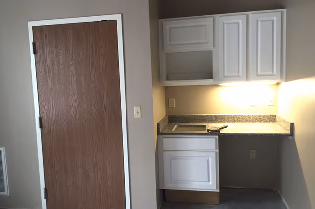 Small kitchenette area with white upper and lower cabinets, a granite countertop with sink, and a closed wooden door to the left.