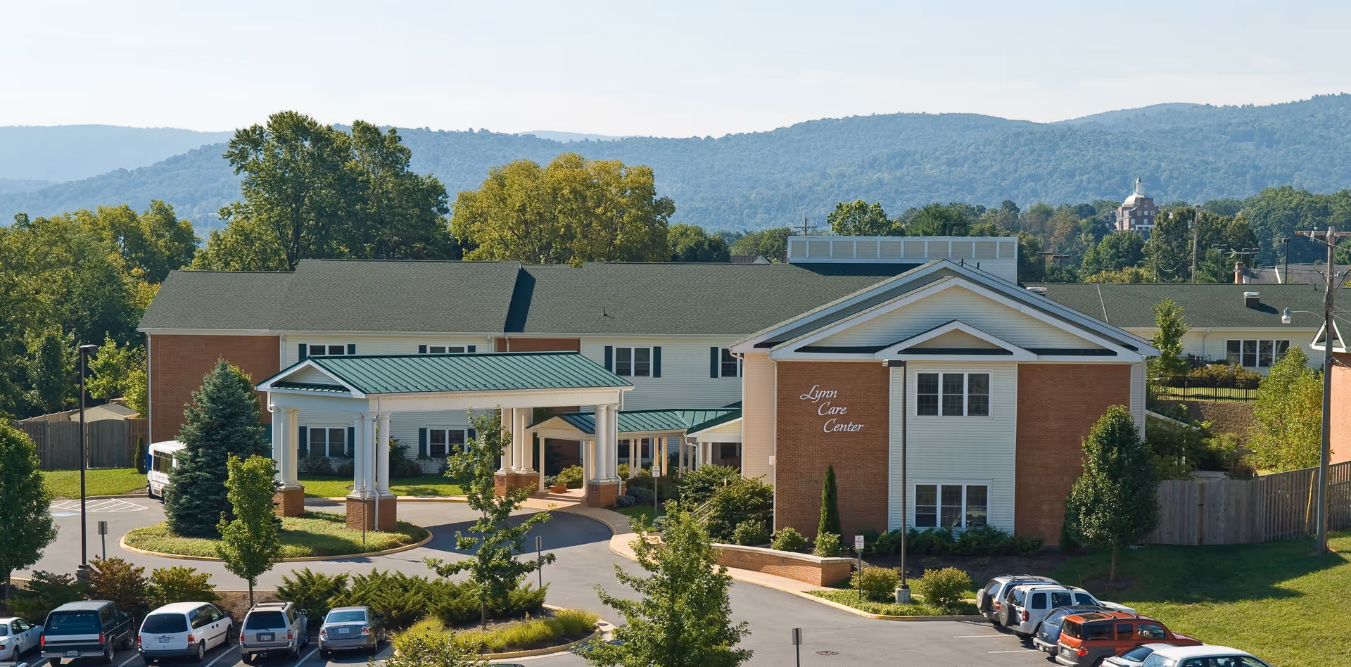 Front view of the Lynn Care Center building with a covered entrance, parking lot, landscaped grounds, and tree-covered hills in the background.