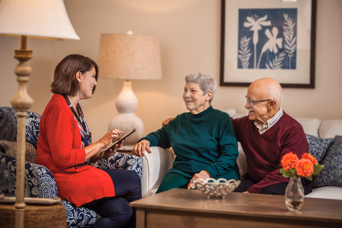 A woman in a red cardigan holding a tablet is sitting on a patterned armchair and talking with an elderly couple seated on a white couch in a cozy living room. The elderly woman is wearing a green sweater and the elderly man is wearing a maroon sweater. There is a wooden coffee table with a decorative bowl and a vase with orange flowers in front of them. A lamp and framed artwork are visible in the background.