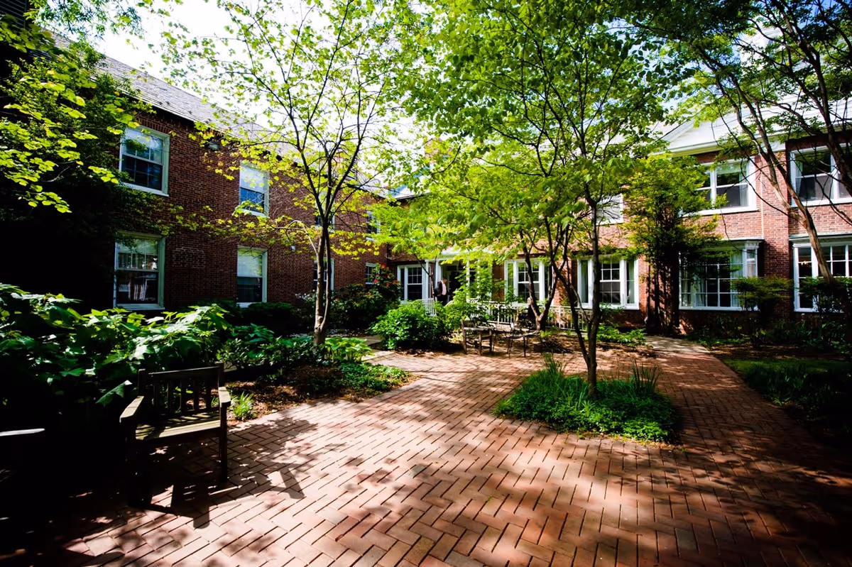 A peaceful outdoor courtyard with brick pathways, surrounded by green trees and shrubs. There are wooden benches and chairs placed along the paths. The courtyard is enclosed by a two-story red brick building with multiple windows.