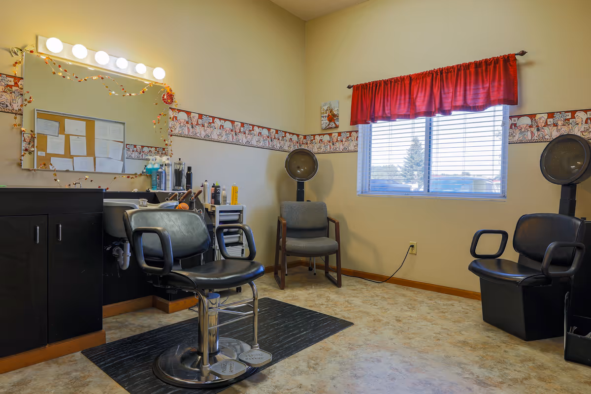 Interior of a small salon room with two black salon chairs, a large mirror with lights above it, various hair care products on a counter, a gray chair, and two hair dryers near a window with red curtains.
