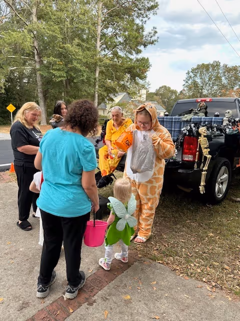 A group of people gathered outdoors near a black pickup truck decorated with Halloween skeletons. One person is dressed in a giraffe costume holding a bag, and a small child dressed as a fairy with green wings is holding a pink bucket. Other adults are standing and sitting nearby, engaging with the child.