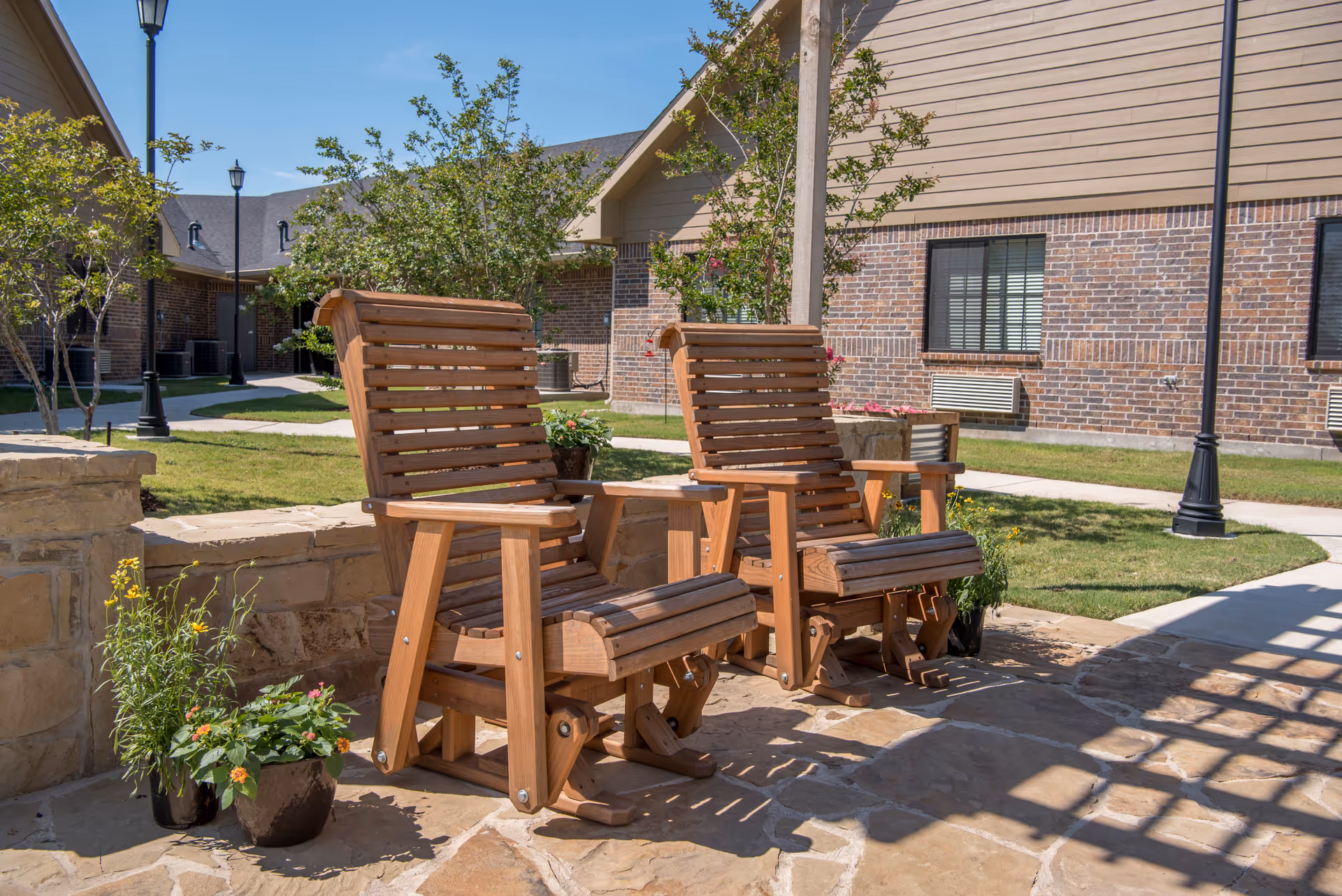 Two wooden rocking chairs on a stone patio with potted plants nearby, set in an outdoor courtyard area with grass, trees, and brick buildings in the background under a clear blue sky.