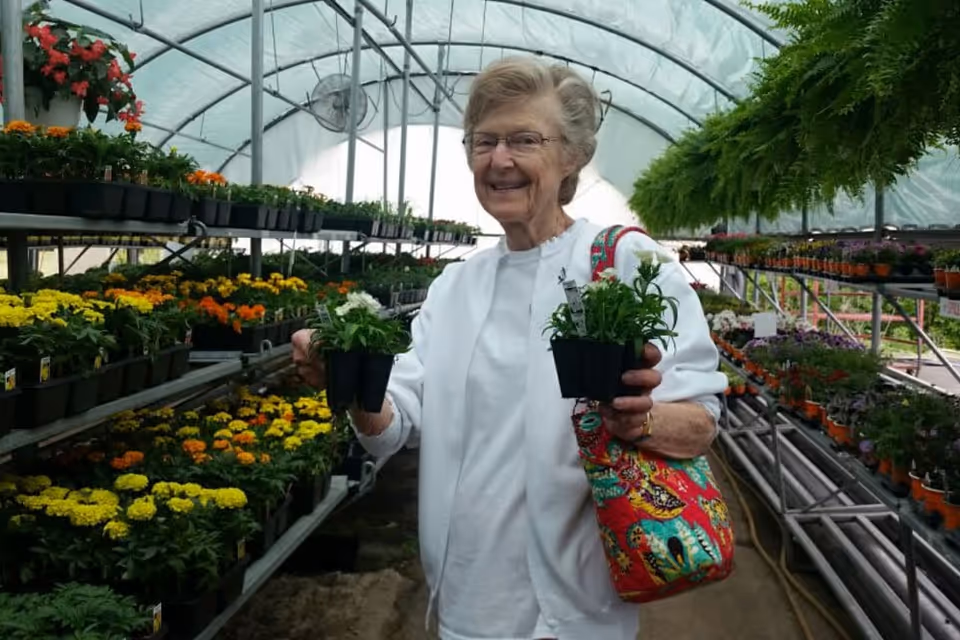 An elderly woman wearing glasses and a white shirt is standing inside a greenhouse filled with various potted flowers and plants. She is smiling and holding two small pots of flowers, with a colorful patterned bag over her shoulder.