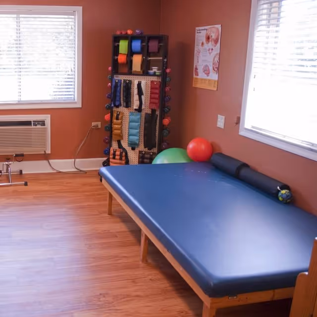 A therapy room with a blue padded treatment table, a black cylindrical bolster, and a small colorful ball on the table. There are two windows with white blinds, an air conditioning unit below one window, and a rack holding various colored therapy bands and weights. The walls are painted a warm brown color, and the floor is wooden.