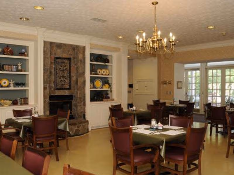 A dining room with several tables covered with green tablecloths and set with placemats and condiments. The room features wooden chairs, built-in white shelves with decorative items, a stone fireplace, and a brass chandelier hanging from the ceiling. Large windows and glass doors allow natural light into the space.