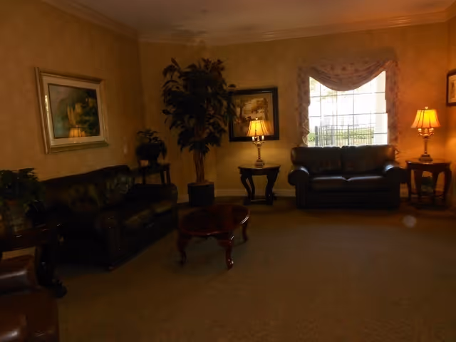 A cozy living room area with two dark leather sofas, a wooden coffee table in the center, two side tables each with a lamp, a large potted plant, framed artwork on the walls, and a window with a decorative valance allowing natural light to enter.