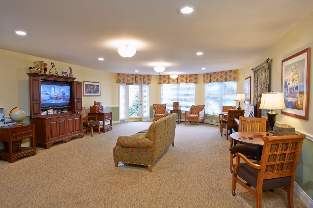 Spacious senior living common room with sofas, armchairs, tables, a TV armoire, and large windows.