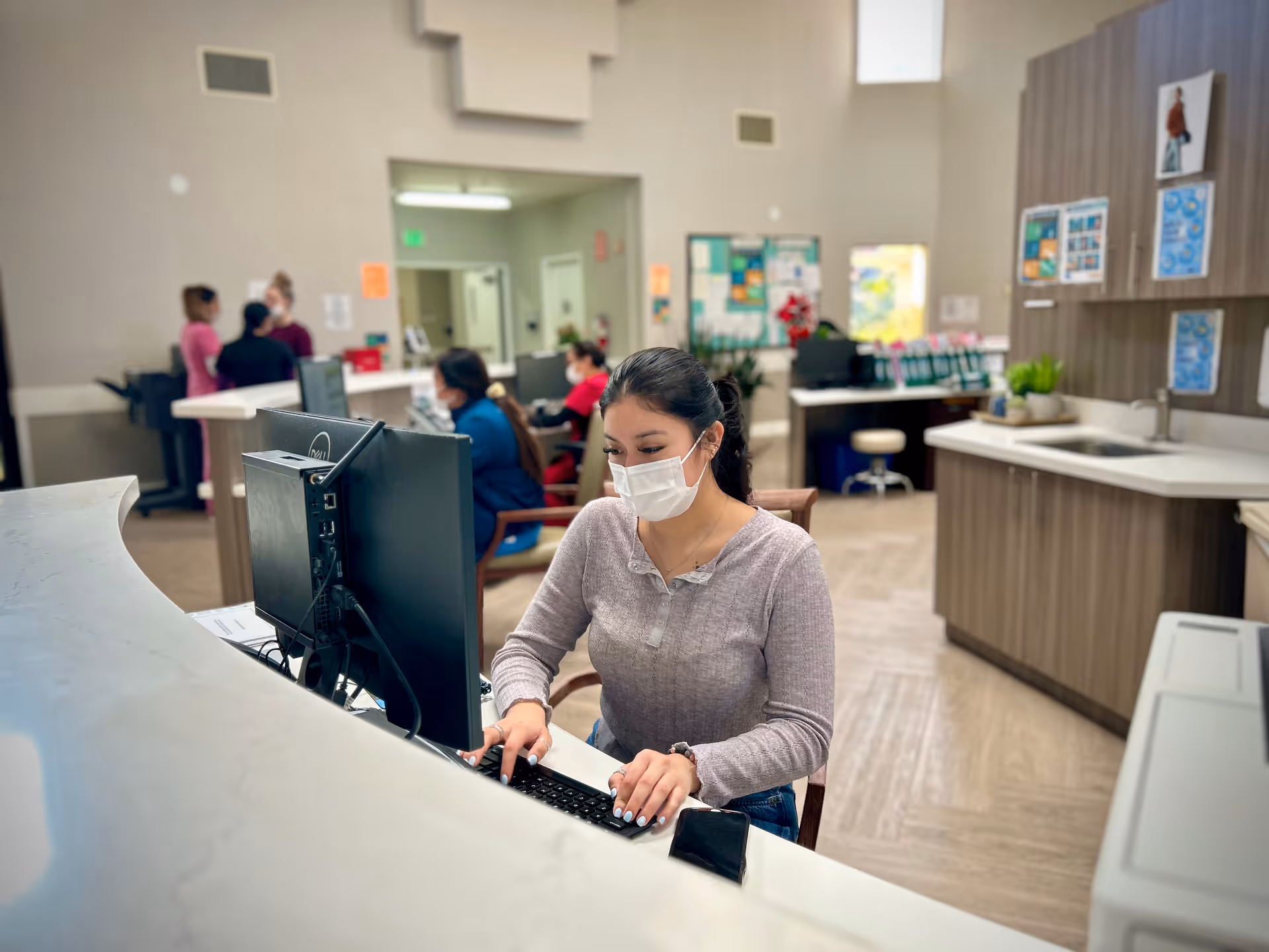 A woman wearing a face mask is seated at a reception desk typing on a keyboard in a well-lit office area. In the background, several other people, also wearing masks, are engaged in conversation or working at computers. The room has light-colored walls, wooden cabinets, and a sink area.