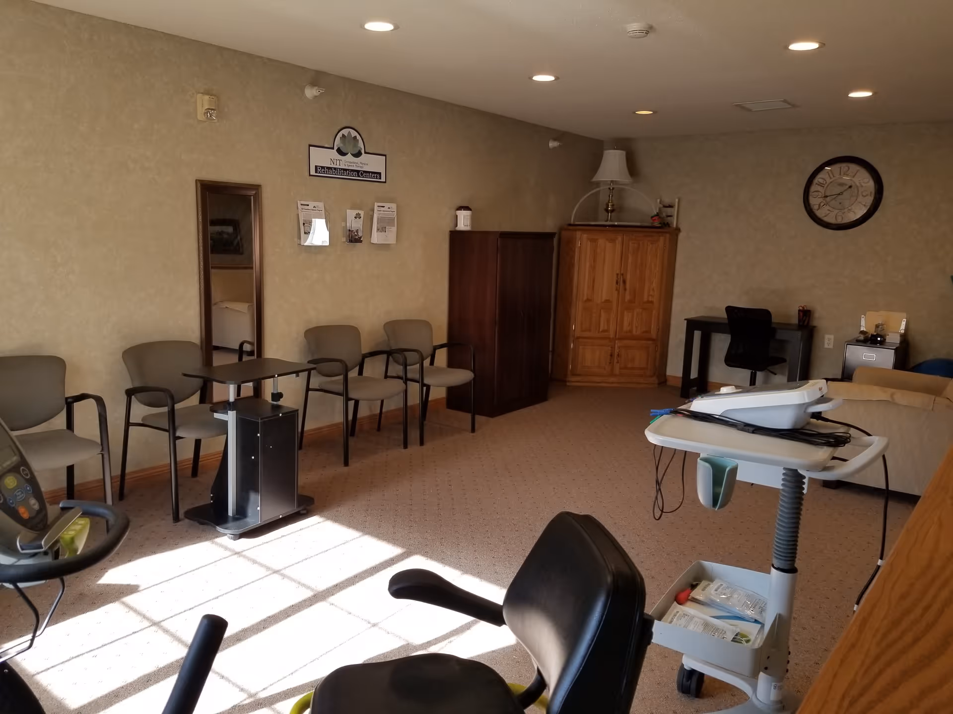 A rehabilitation center room with exercise equipment including a stationary bike, several chairs lined up against the wall, a small table, a wooden cabinet, a desk with a chair, a clock on the wall, and a lamp on top of a cabinet. The room is carpeted and well-lit with ceiling lights.
