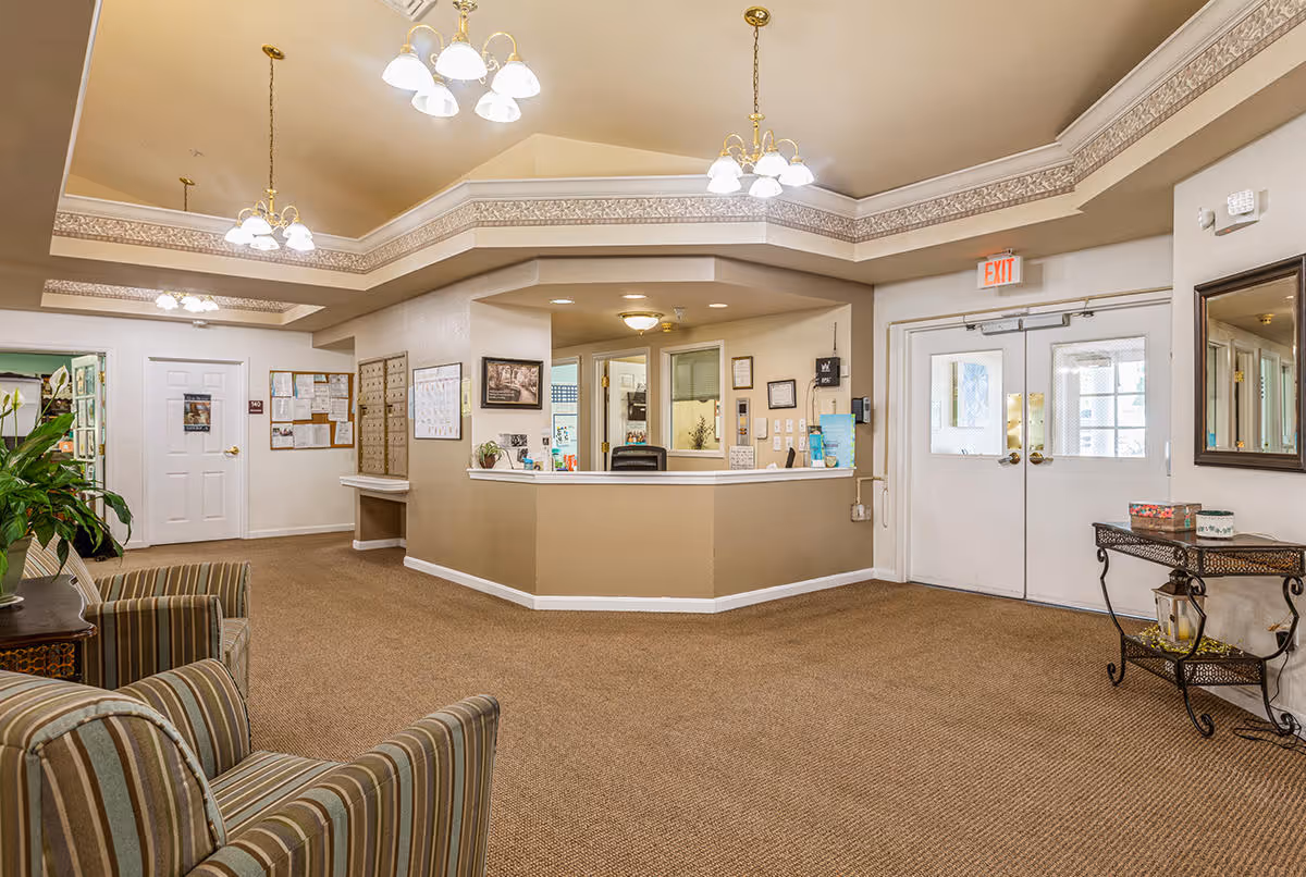 Reception area of The Addison of Lowrie Place featuring a beige front desk with office supplies, a bulletin board with notices, striped armchairs, a small decorative table with items on it, and double doors with an exit sign above. The ceiling has decorative molding and multiple hanging light fixtures.