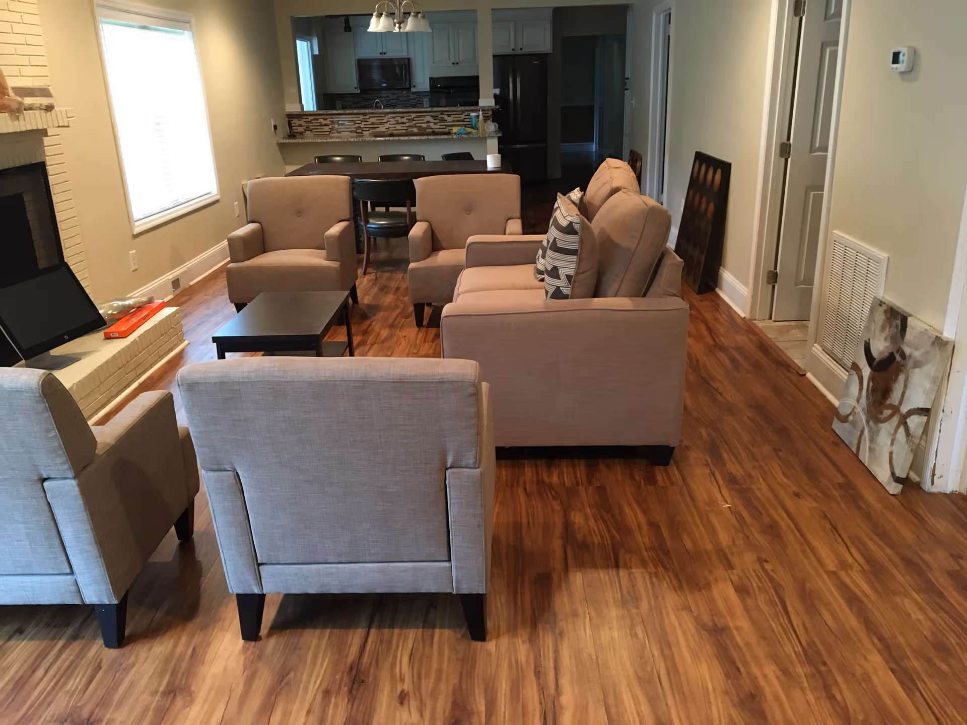 A living room area with beige and light gray armchairs and a sofa arranged around a small black coffee table. The room has wooden flooring, a white brick fireplace on the left, and a window with blinds. In the background, there is a kitchen area with a counter and bar stools, and some artwork leaning against the wall on the right side.