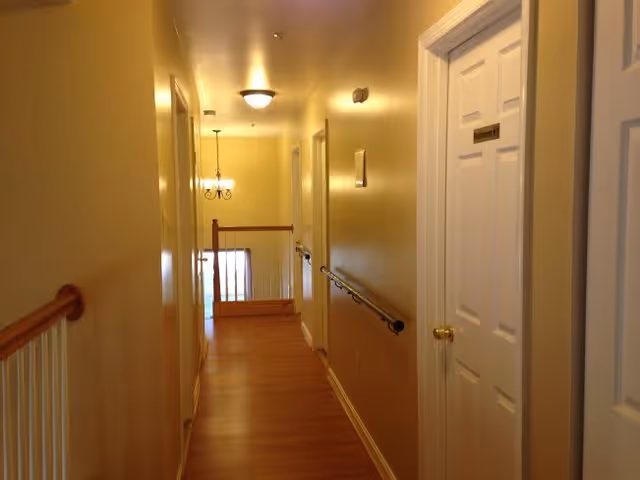 A well-lit indoor hallway with wooden flooring and beige walls. There are white doors on the right side, one labeled 'Restroom'. A handrail runs along the right wall, and a wooden railing is visible at the end of the hallway near a hanging light fixture.