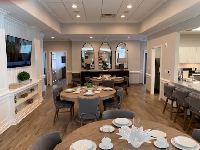 Interior view of a senior care facility dining area with round wooden tables set with white plates, cups, and glassware. There are gray upholstered chairs around the tables. The room has wood flooring, a wall-mounted TV, decorative mirrors, and a kitchen counter with bar stools on the right side.