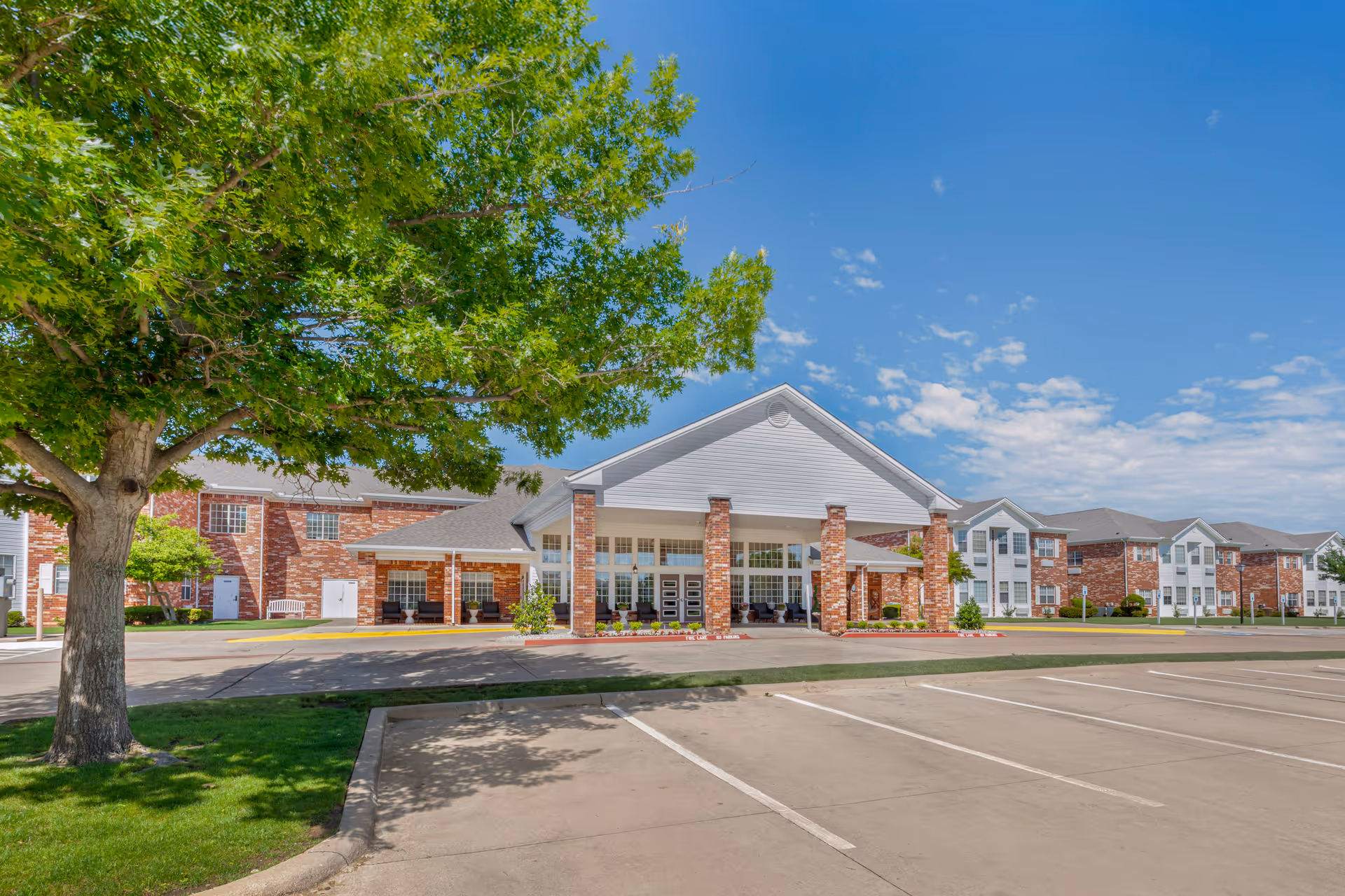 Front entrance of a red-brick senior living facility with a covered portico, parking lot, and a large tree in the foreground.
